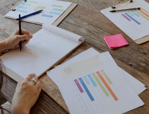 Hands writing on paper with colorful charts on wooden table, organized workspace.